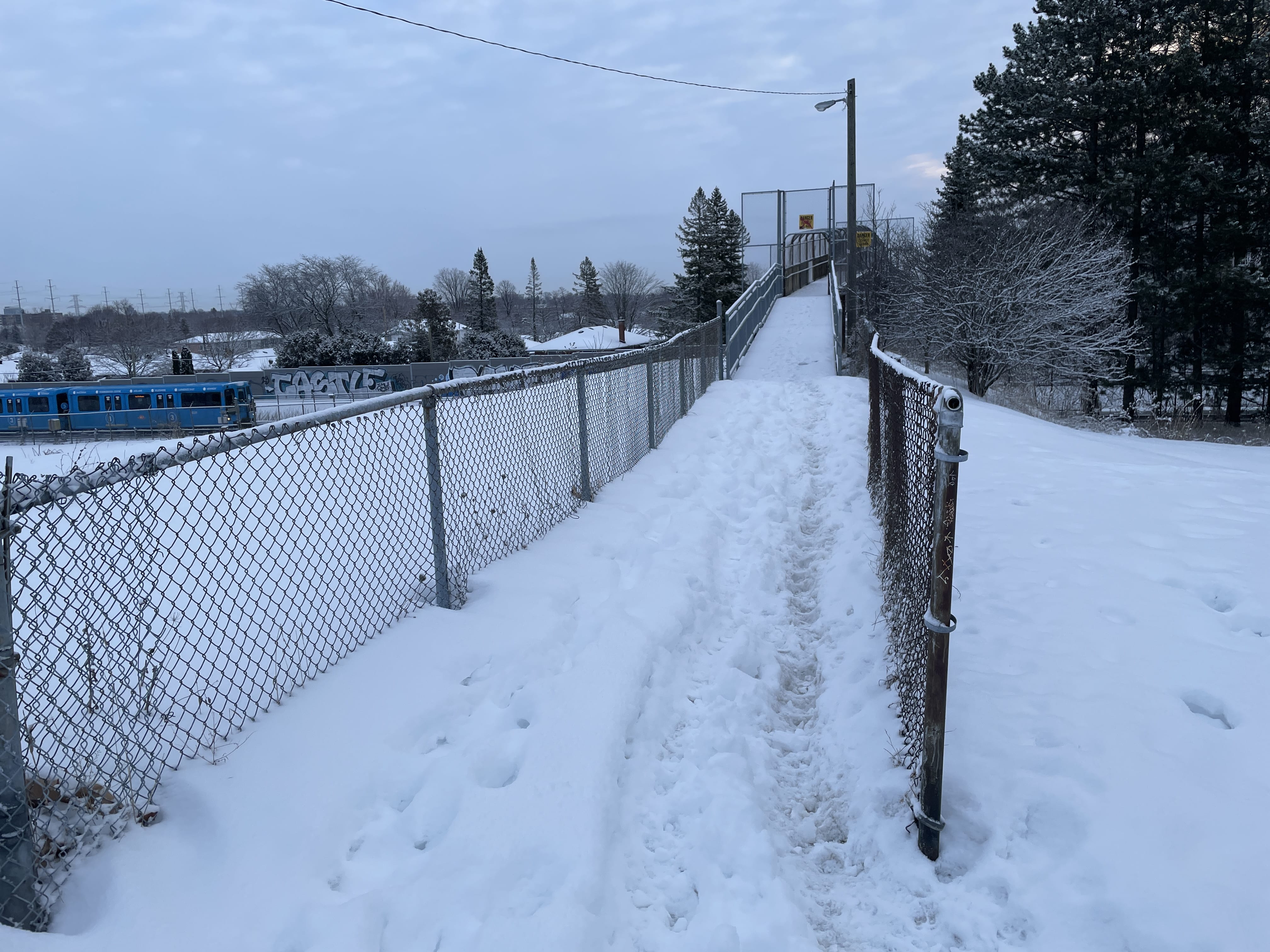 Daytime photo of the pedestrian bridge over the RT line and the narrow path leading up to it on the west side. The path is covered in snow that is at least 15-30cm deep. A blue line 3 train is visible in the background, passing under the bridge.